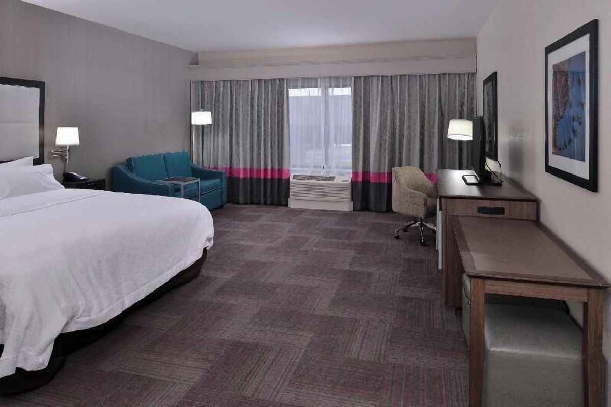 A hotel room with a bed, a desk, a chair, a blue armchair, and a large window with curtains.