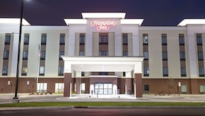 Un hotel Hampton Inn con una fachada moderna, una entrada central y un asta con la bandera estadounidense.