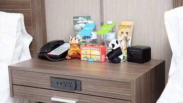 A hotel room with a wooden nightstand holding a telephone, a Barnum's Animals box, a panda plush toy, a water bottle, and a book titled "Explode".