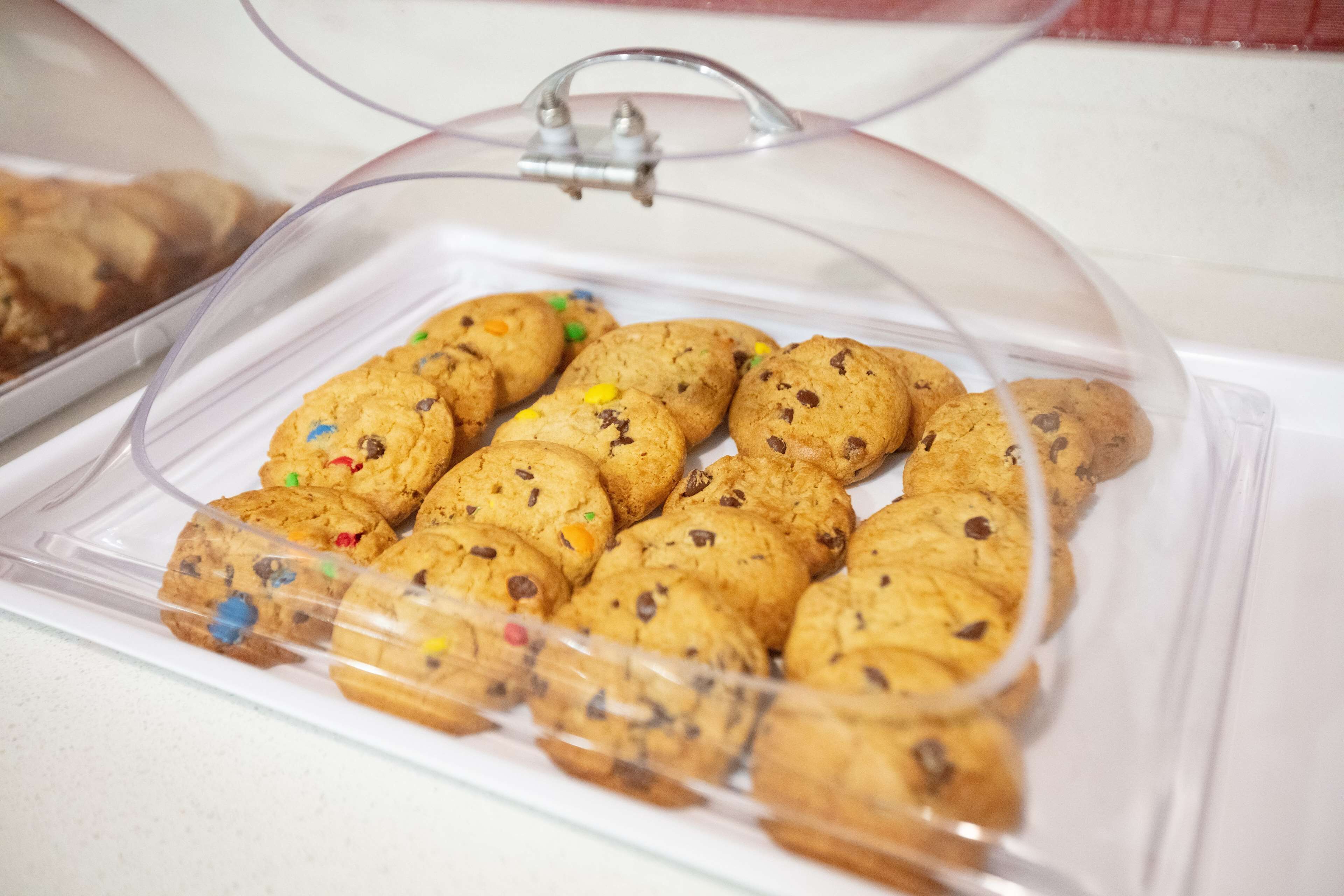 A clear plastic container with a glass dome lid, holding a variety of cookies with visible chocolate chips and colorful sprinkles.