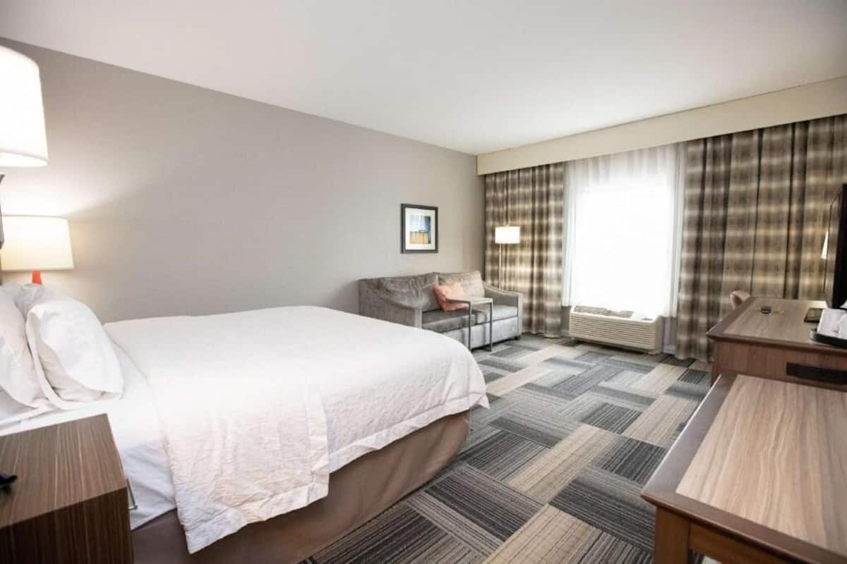 Quarto de hotel com cama, escrivaninha, cadeira, luminária, quadro na parede e uma janela grande com cortinas.
