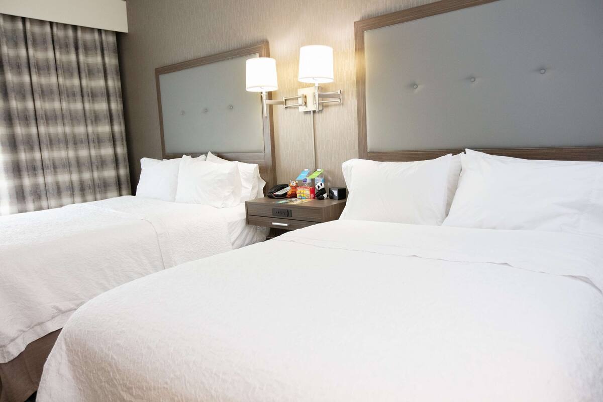 Quarto de hotel com duas camas, uma mesinha de cabeceira e uma luminária.