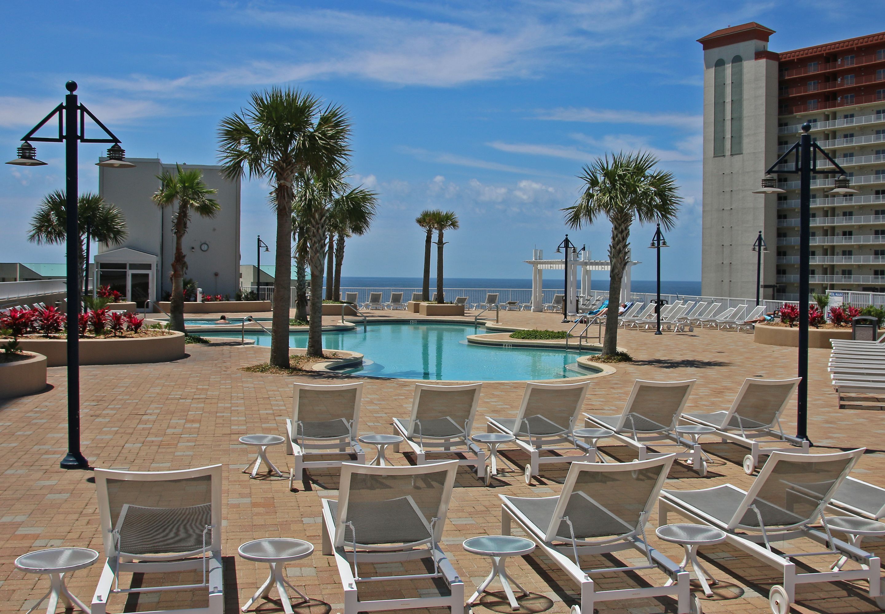 A poolside area with lounge chairs and a pool, surrounded by palm trees and a building with a red roof.