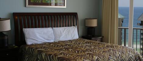 A hotel room with a large bed, two bedside tables with lamps, a framed artwork above the bed, and a view of the ocean through a balcony.