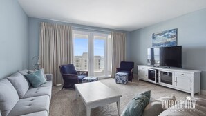 Una sala de estar moderna con un sofá, sillones, una mesa de centro y un televisor sobre un mueble blanco. Amplias ventanas con cortinas ofrecen vistas de la ciudad.