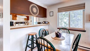 A dining area with a wooden table and four chairs, a kitchen with wooden cabinets and a built-in oven, and a large window with a view of trees. - Mark IX 1 By Peak Property Management (Breckenridge)
