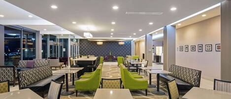 A modern hotel lobby with a central seating area, multiple tables and chairs, and large windows offering a view of the outside.
