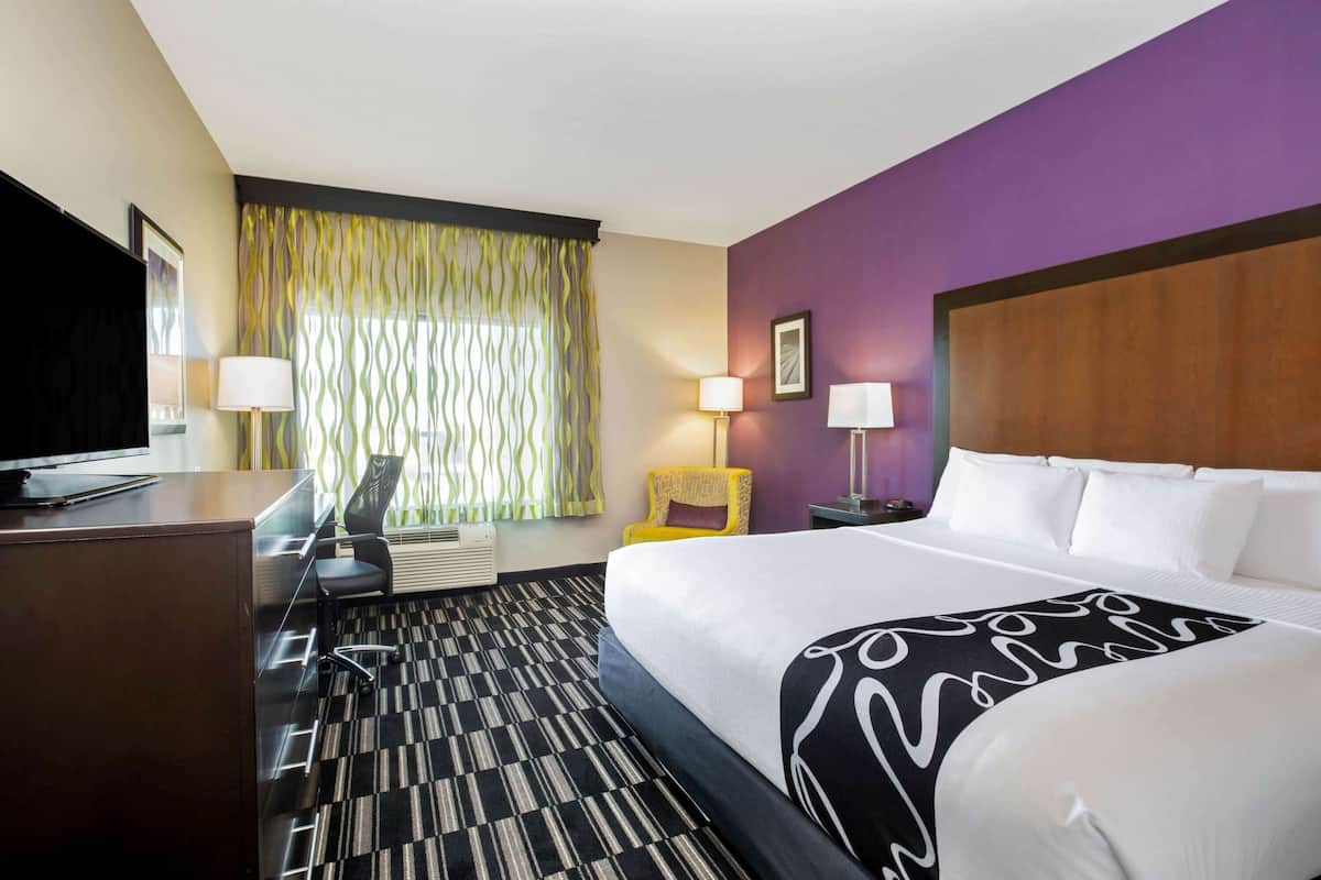 Habitación de hotel con una cama grande, un escritorio con televisor, una silla y un ventanal con cortinas.