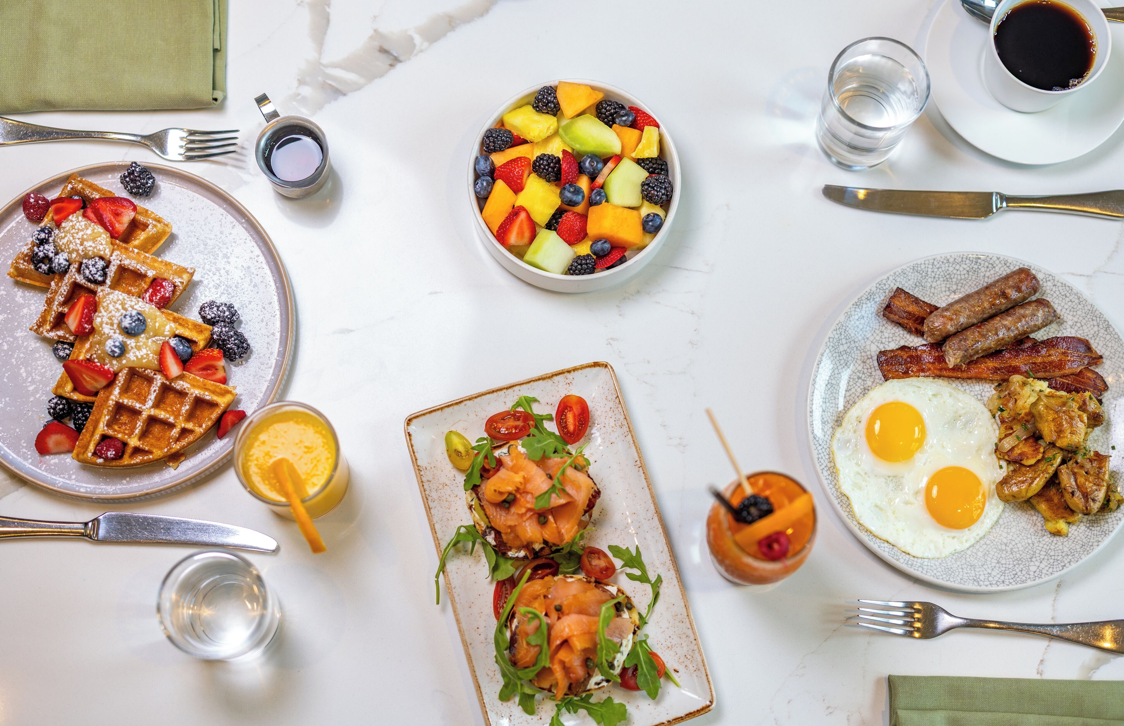 A breakfast buffet with waffles, fruit salad, smoked salmon, eggs, and bacon.