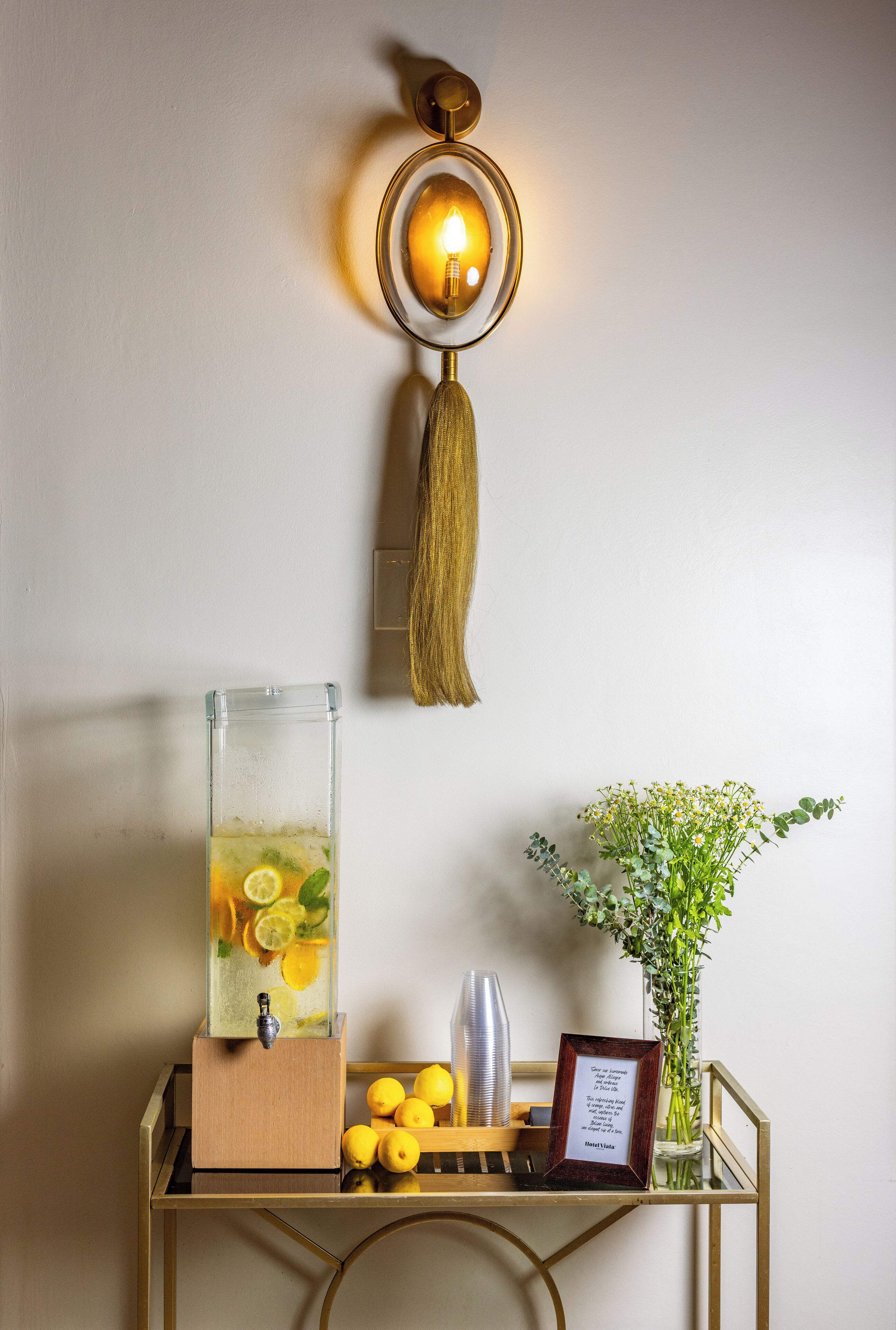 A wall-mounted light fixture with a tassel, a glass pitcher with citrus slices, lemons, and a framed quote on a metal stand.