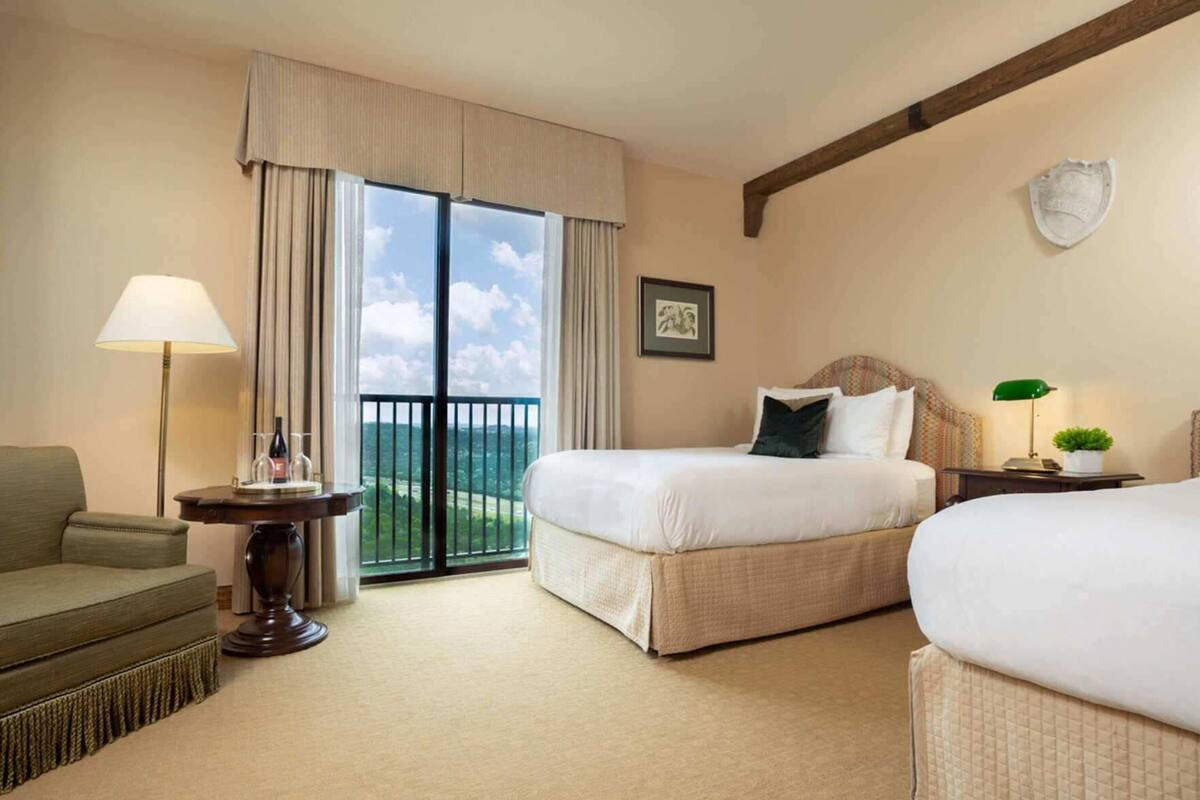 A hotel room with a bed, a chair, a table with a lamp, a small plant, and a balcony with a view.
