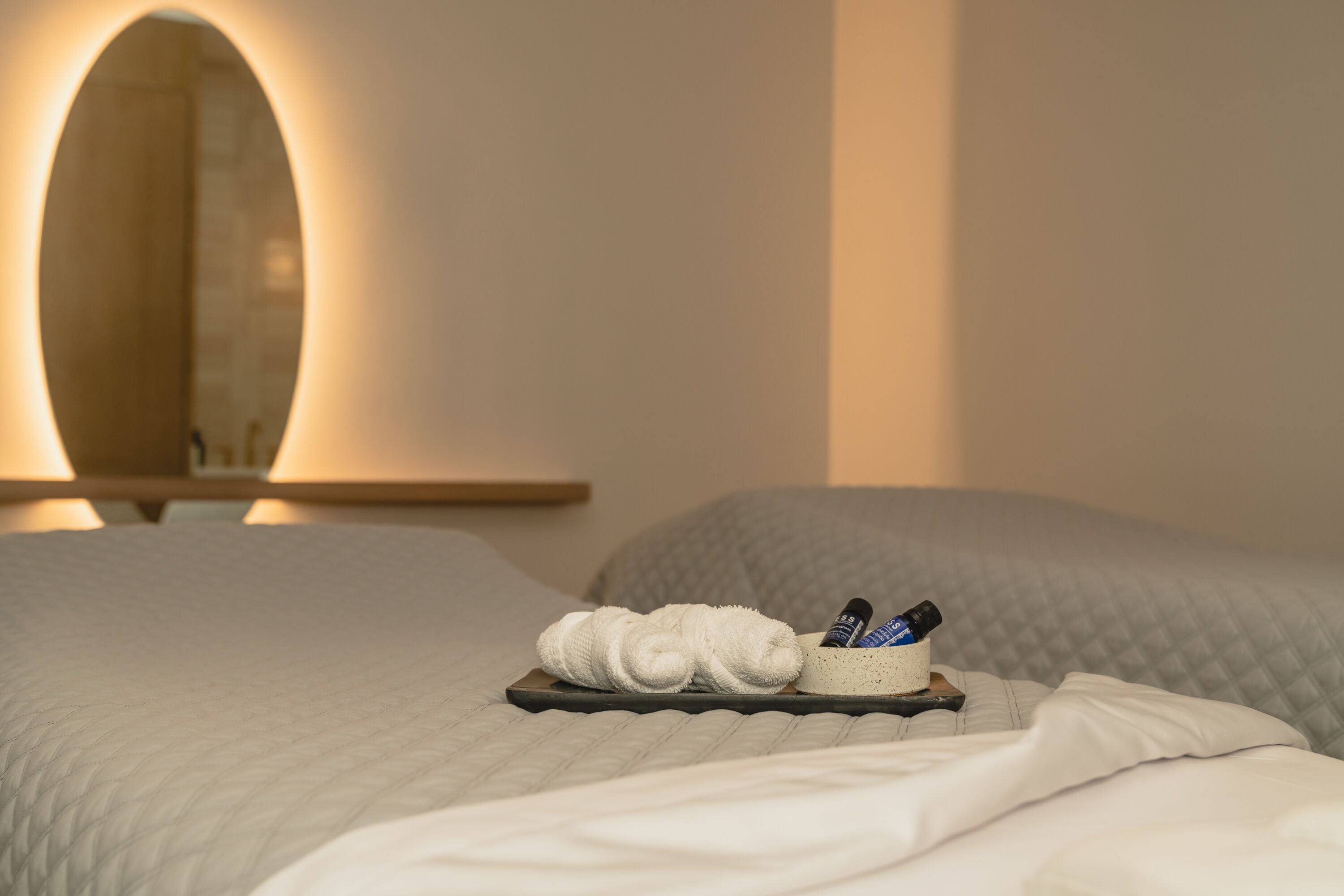 Un lit bien fait, avec un plateau contenant des articles de toilette, notamment une serviette et des flacons, dans une chambre d’hôtel dotée d’un grand miroir et d’une table de chevet en bois.