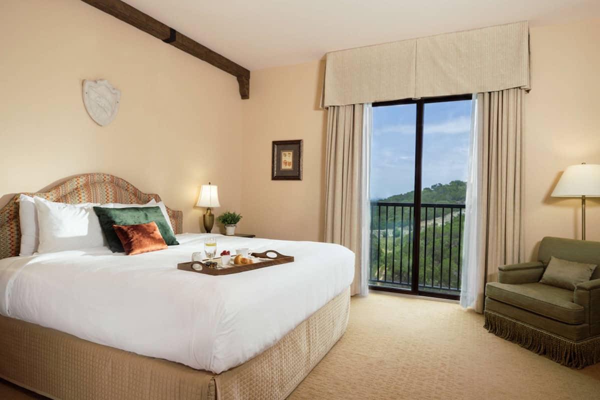 A hotel room with a large bed, a balcony with a view, a green armchair, and a tray of breakfast items on the bed.