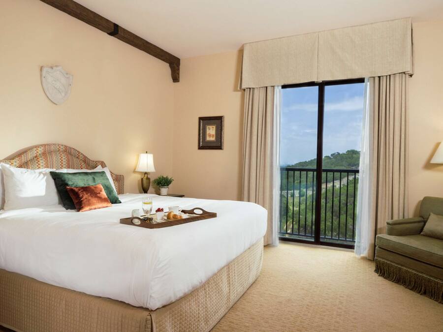 A hotel room with a large bed, a balcony with a view, a green armchair, and a tray of breakfast items on the bed.