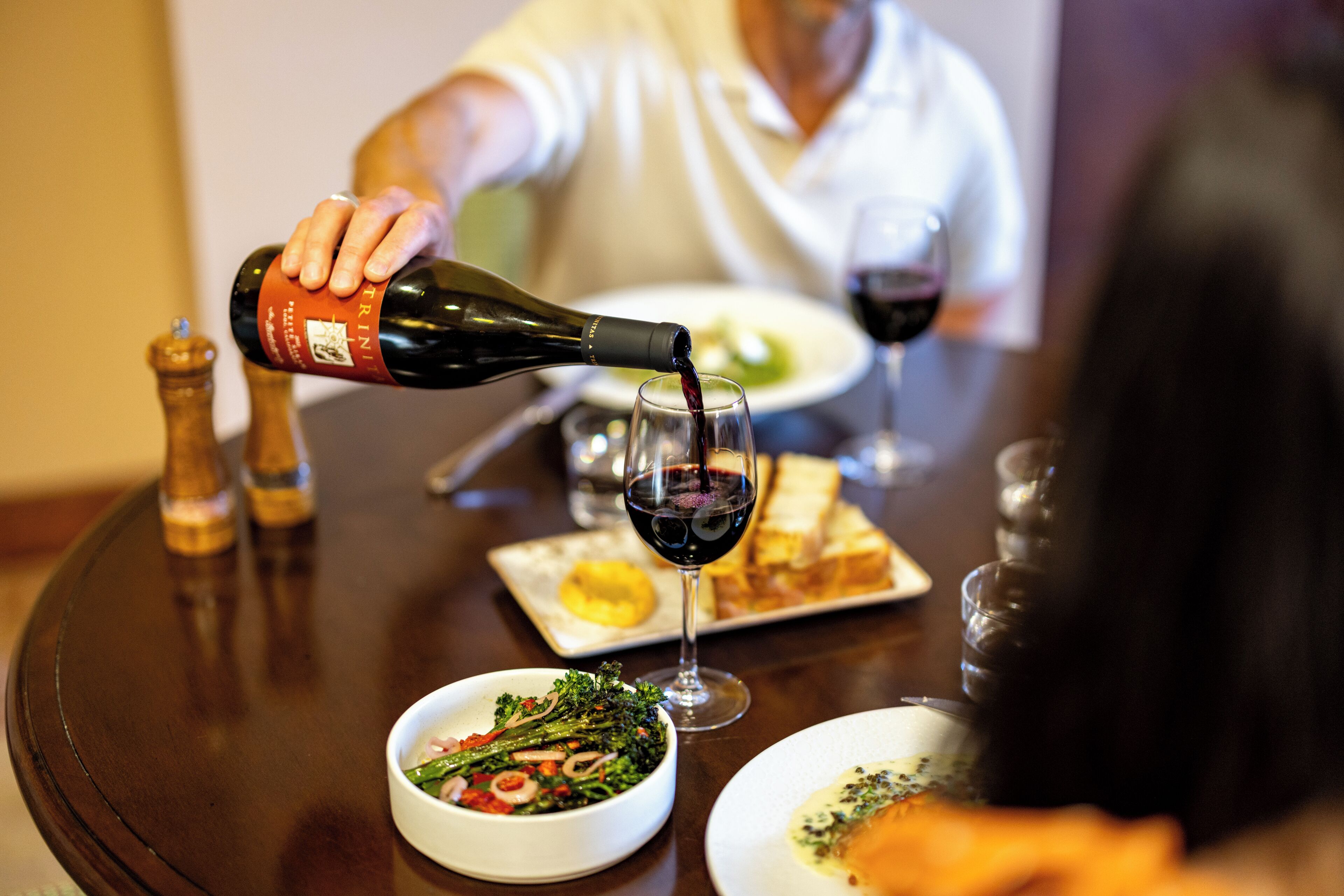 Una mesa de comedor con una botella de vino tinto sirviéndose en una copa, un tazón de ensalada y platos de comida.