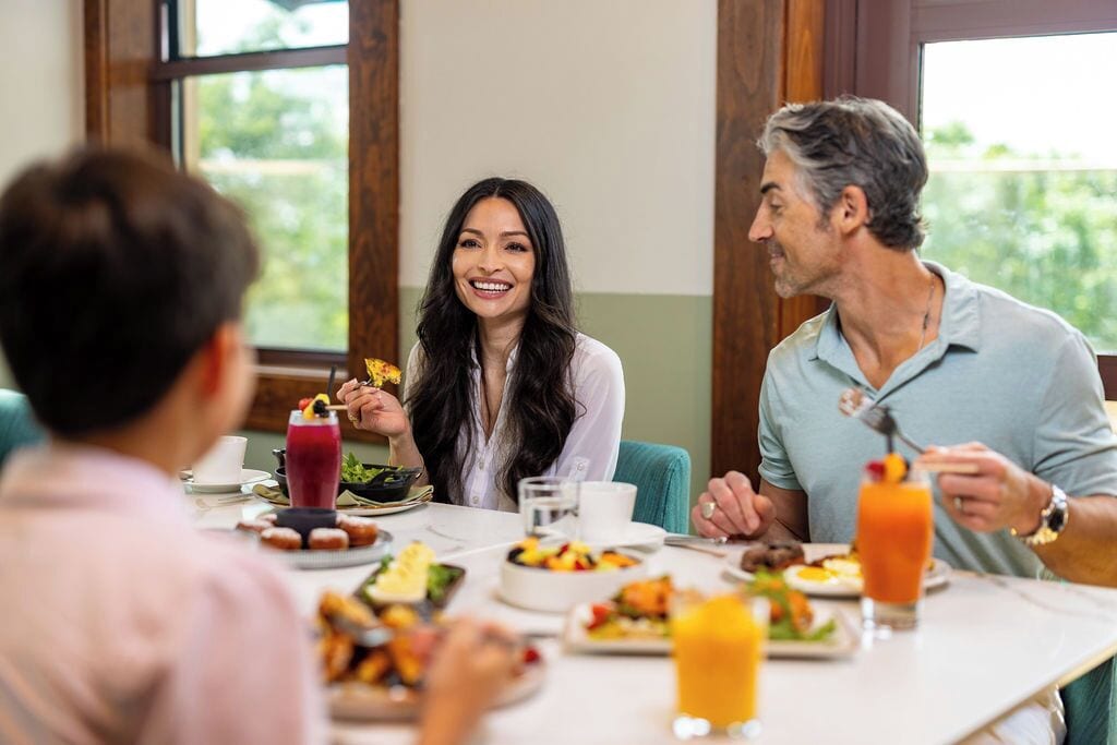 A dining table with various dishes and drinks, a window with a view of greenery, and a person in a light blue shirt.