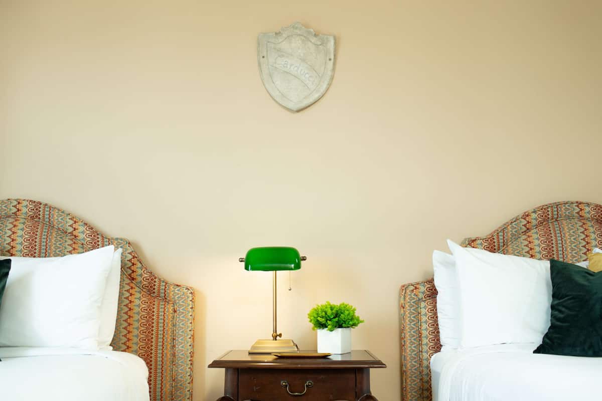 A hotel room with two beds, patterned headboards, a nightstand with a green desk lamp, a small potted plant, and a decorative shield on the wall.