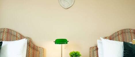 A hotel room with two beds, patterned headboards, a nightstand with a green desk lamp, a small potted plant, and a decorative shield on the wall.