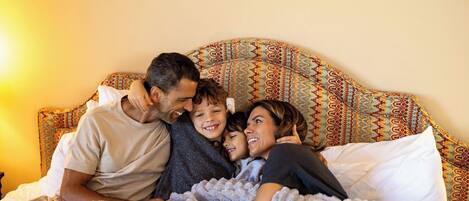 A family of four in a bedroom with a patterned headboard, a bed with white linens, and a blanket.