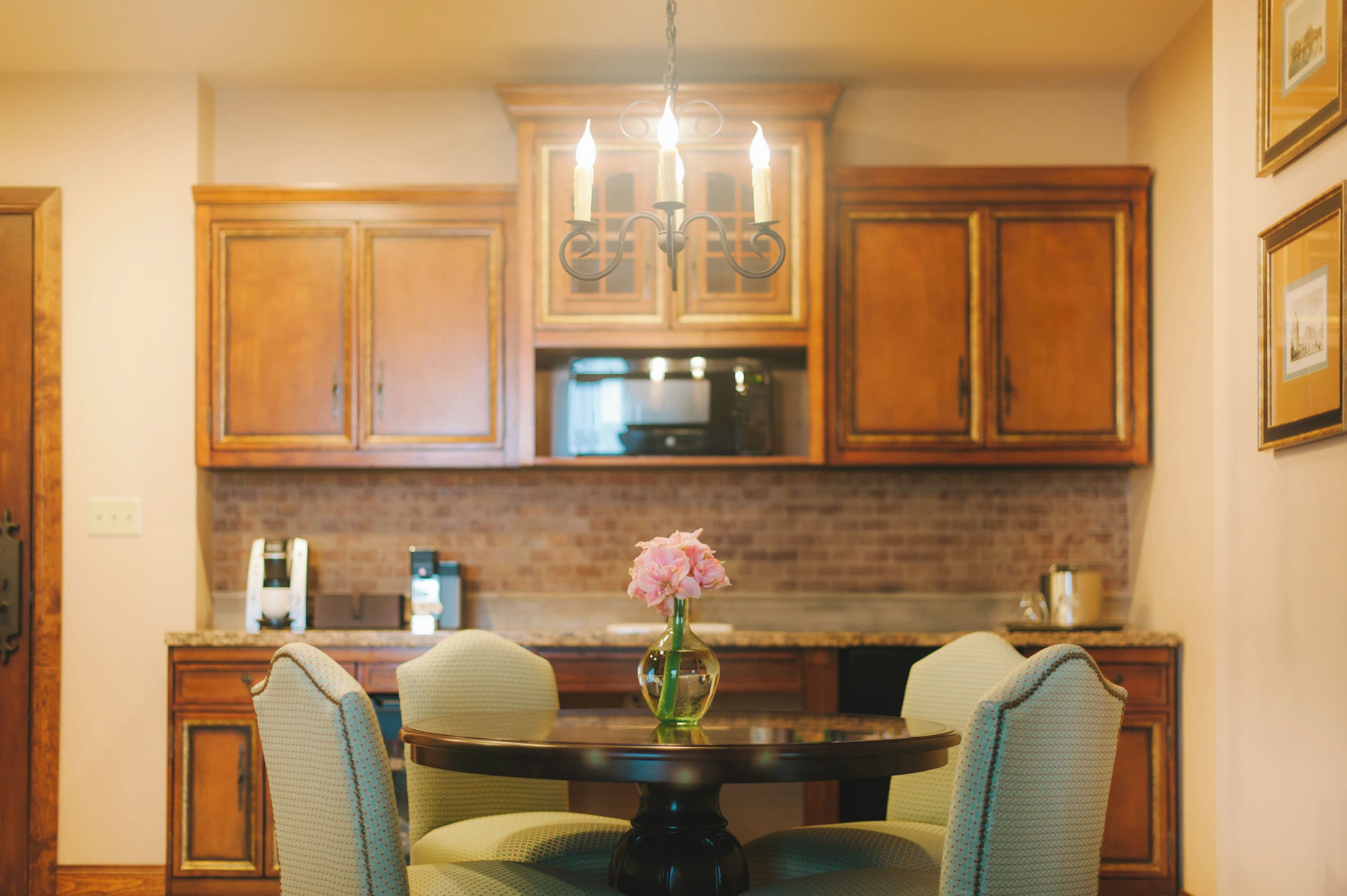 A kitchen with wooden cabinets, a chandelier, a round table with chairs, and a vase with flowers.