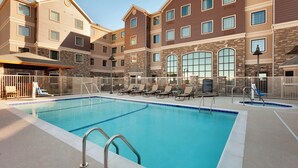 A swimming pool with a white tiled edge, surrounded by a white fence, and adjacent to a multi-story building with a brick facade and multiple windows. - Staybridge Suites Midland by IHG (Midland)