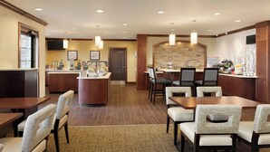 A hotel dining area with a central island, seating arrangements, and a kitchen in the background.