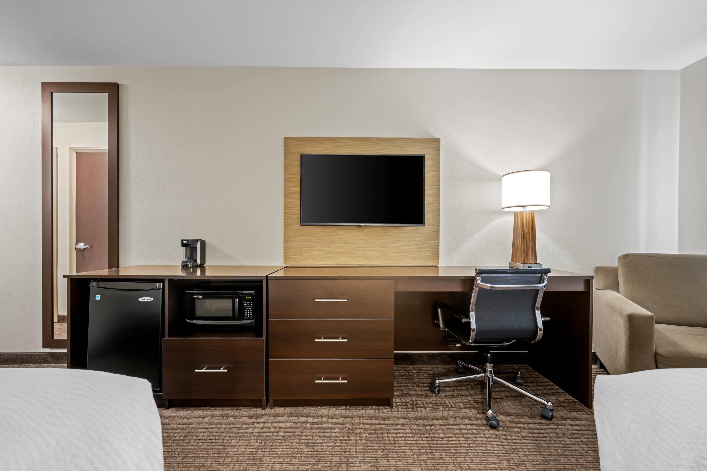 A hotel room with a desk, a flat-screen TV, a lamp, a microwave, a mini-fridge, a chair, and two beds.