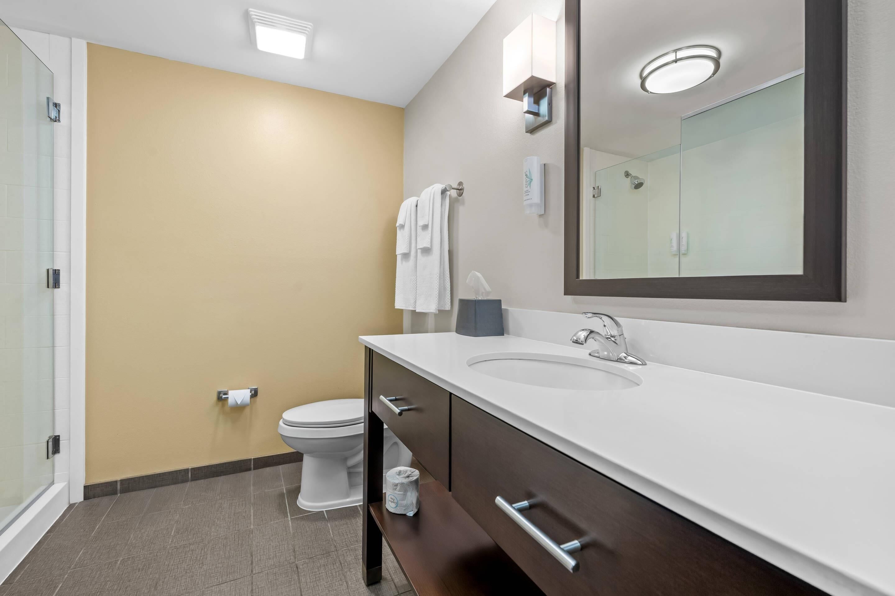 Een moderne badkamer met een behang in beige tint, een wit toilet, een grote spiegel en een witte wastafel met een donkere kast.