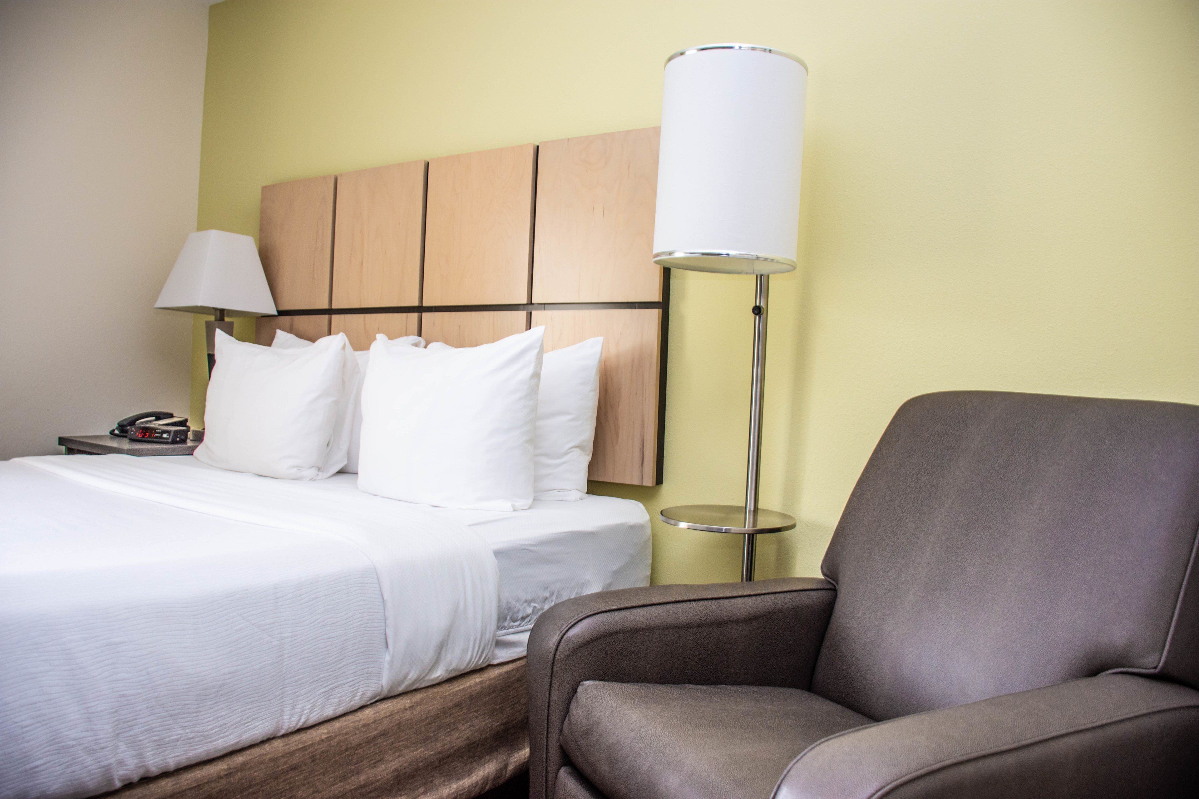 A hotel room with a bed, a chair, a bedside table with a lamp, and a wall-mounted telephone.
