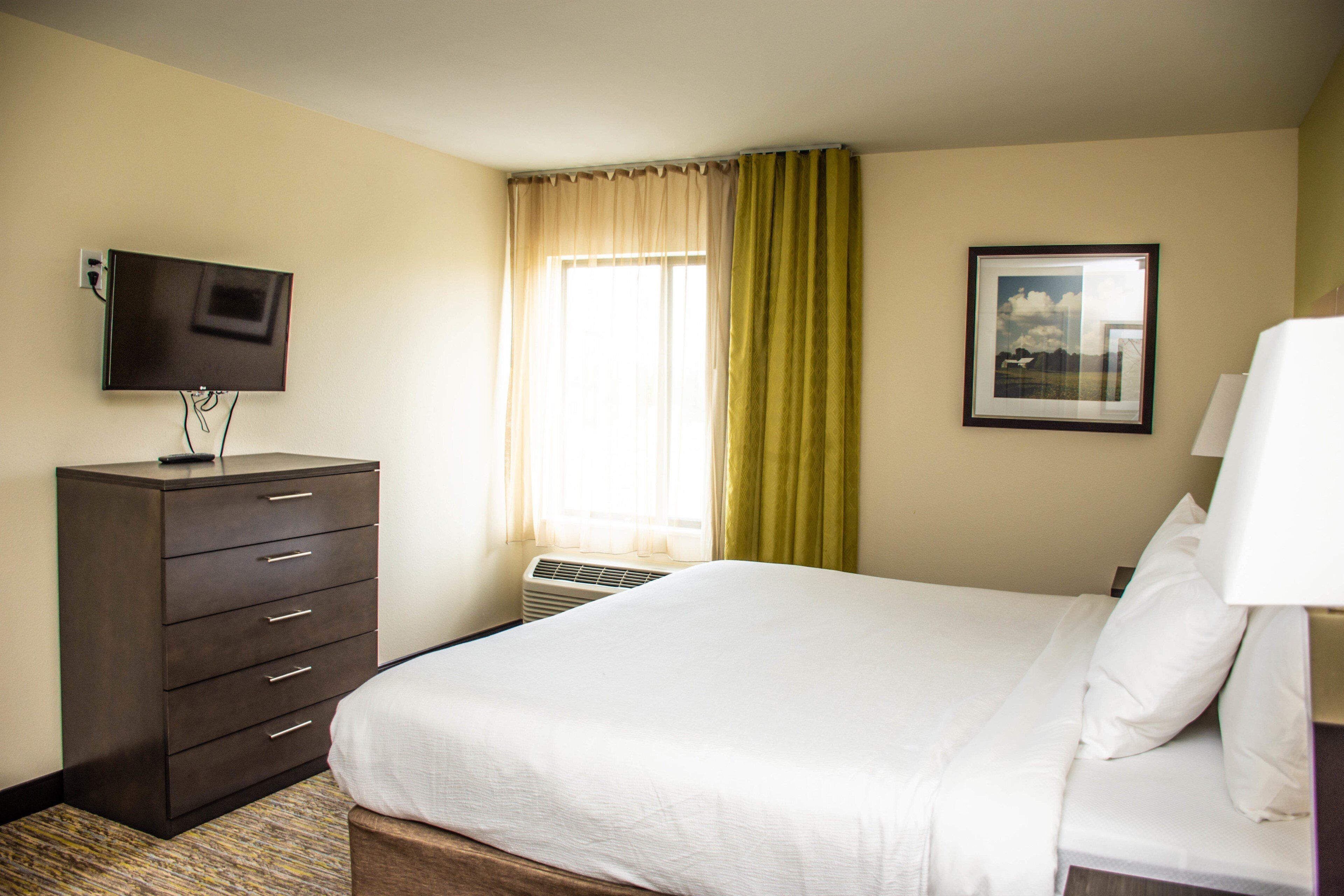 A hotel room with a bed, a dresser, a television, a window with curtains, and a framed picture on the wall.
