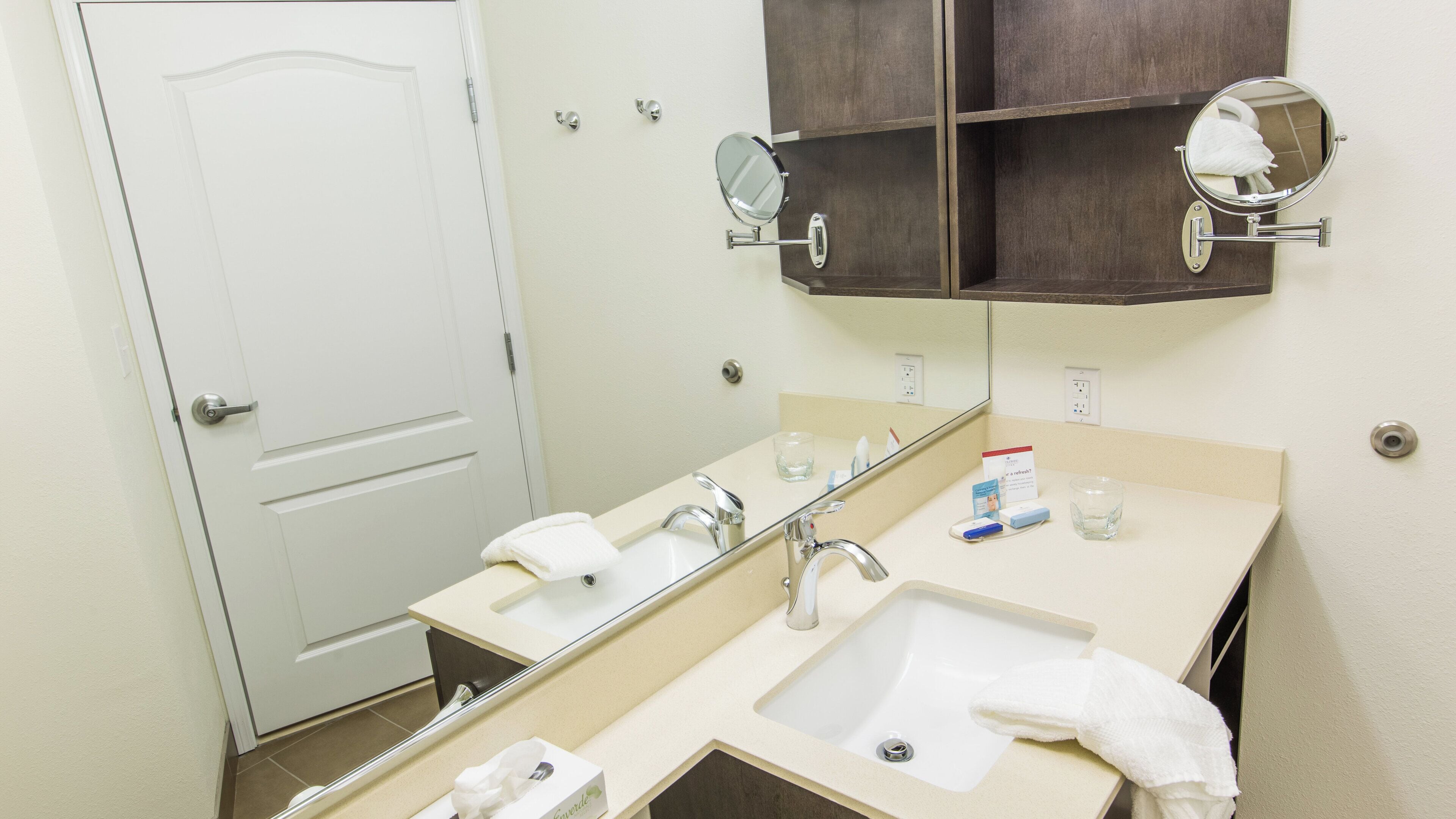 Un baño de hotel con un lavamanos blanco, dos espejos, una toalla y un vaso de agua.
