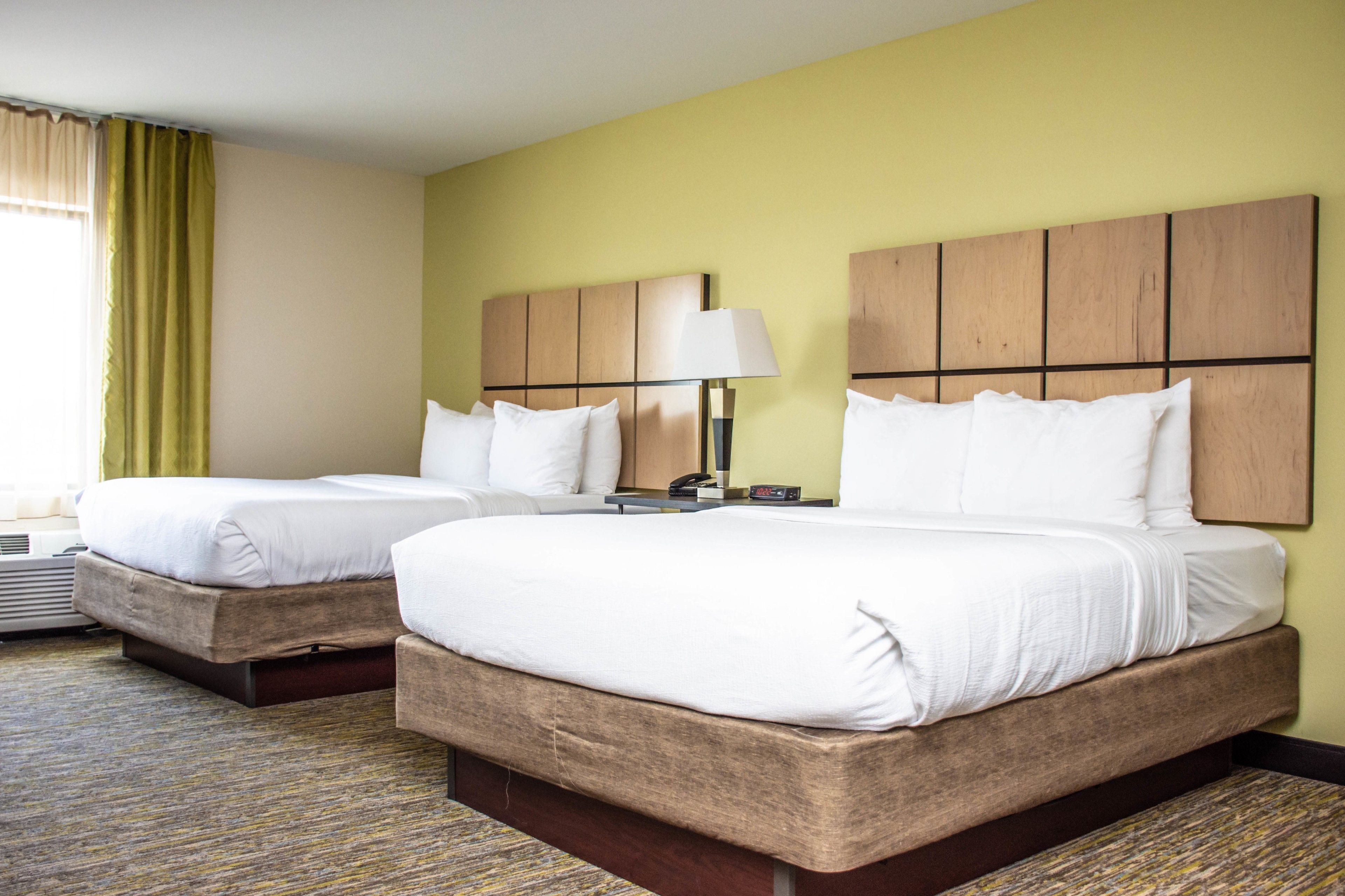 A hotel room with two beds, a bedside table with a lamp, and a large window with curtains.