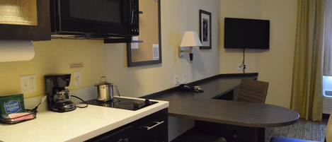 A compact hotel room with a kitchenette, a flat-screen TV mounted on the wall, a small table, and a single chair.