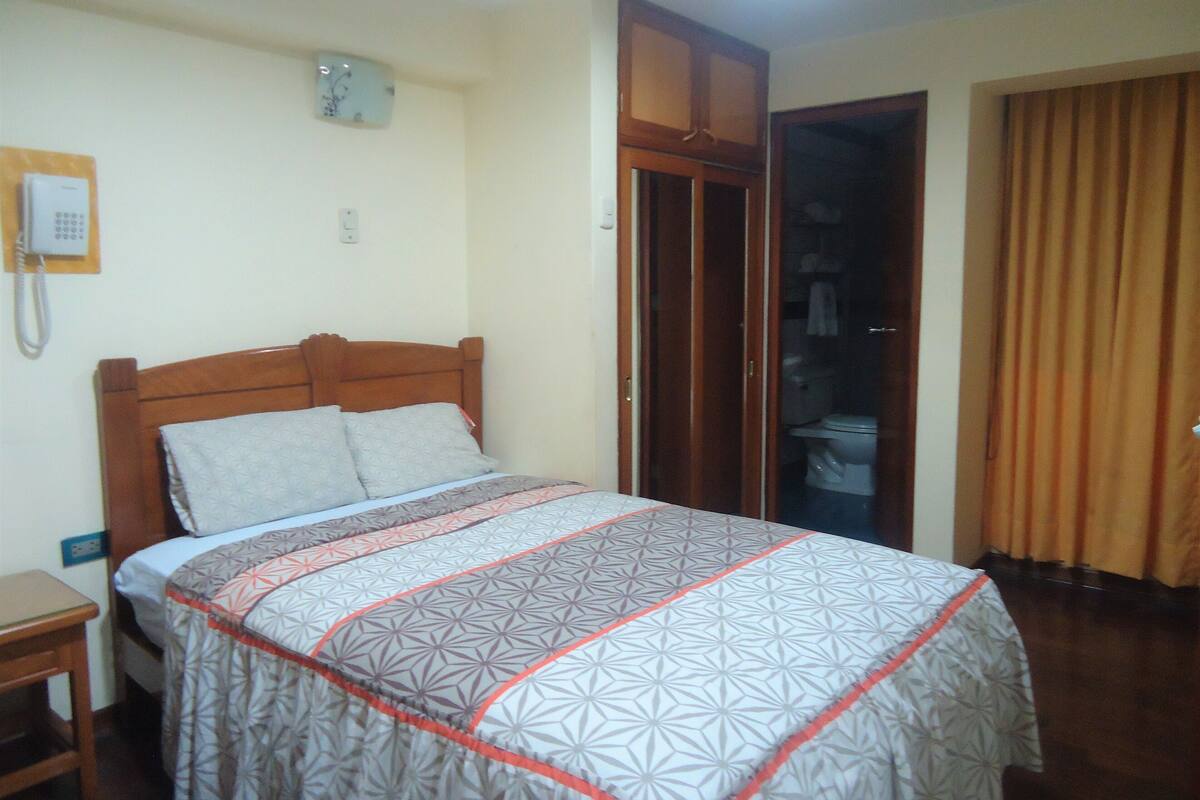 A bedroom with a bed, a bedside table, a wardrobe, and a bathroom visible through an open door.