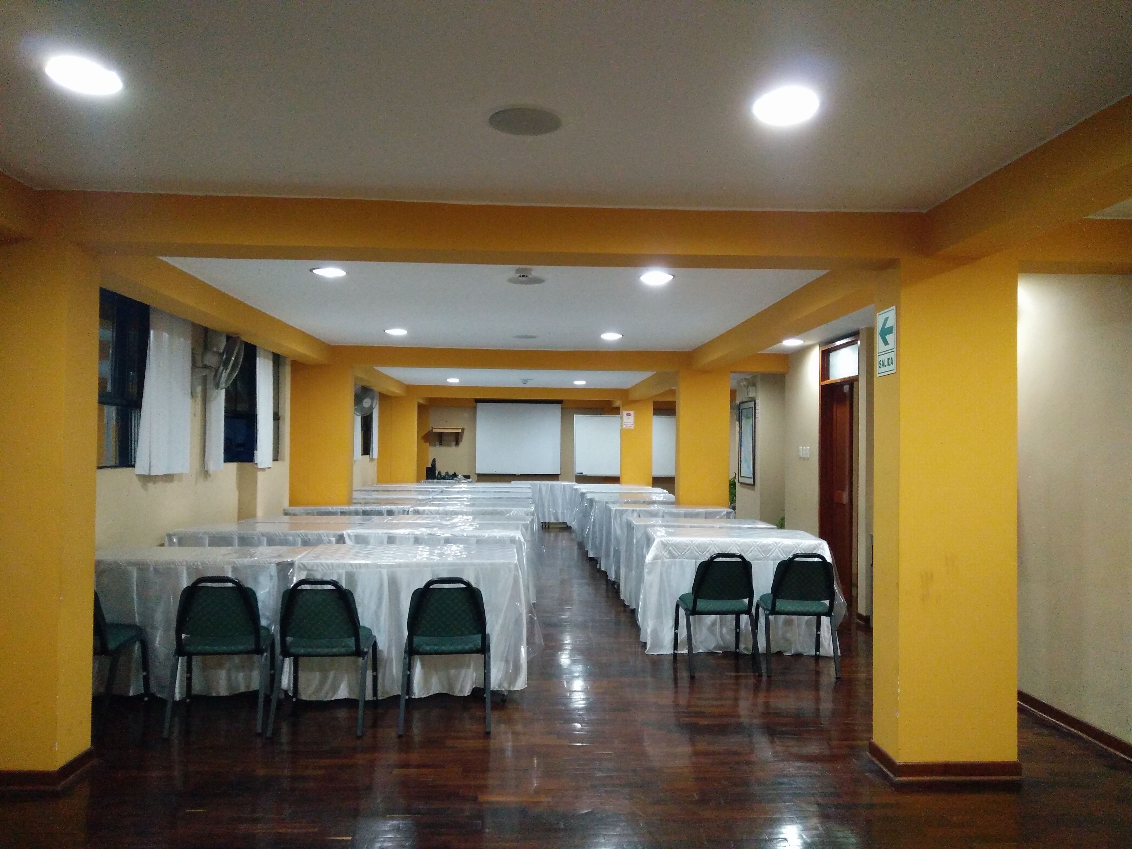 Un salón de conferencias con filas de mesas y sillas, una pantalla de proyector y una señal de salida.