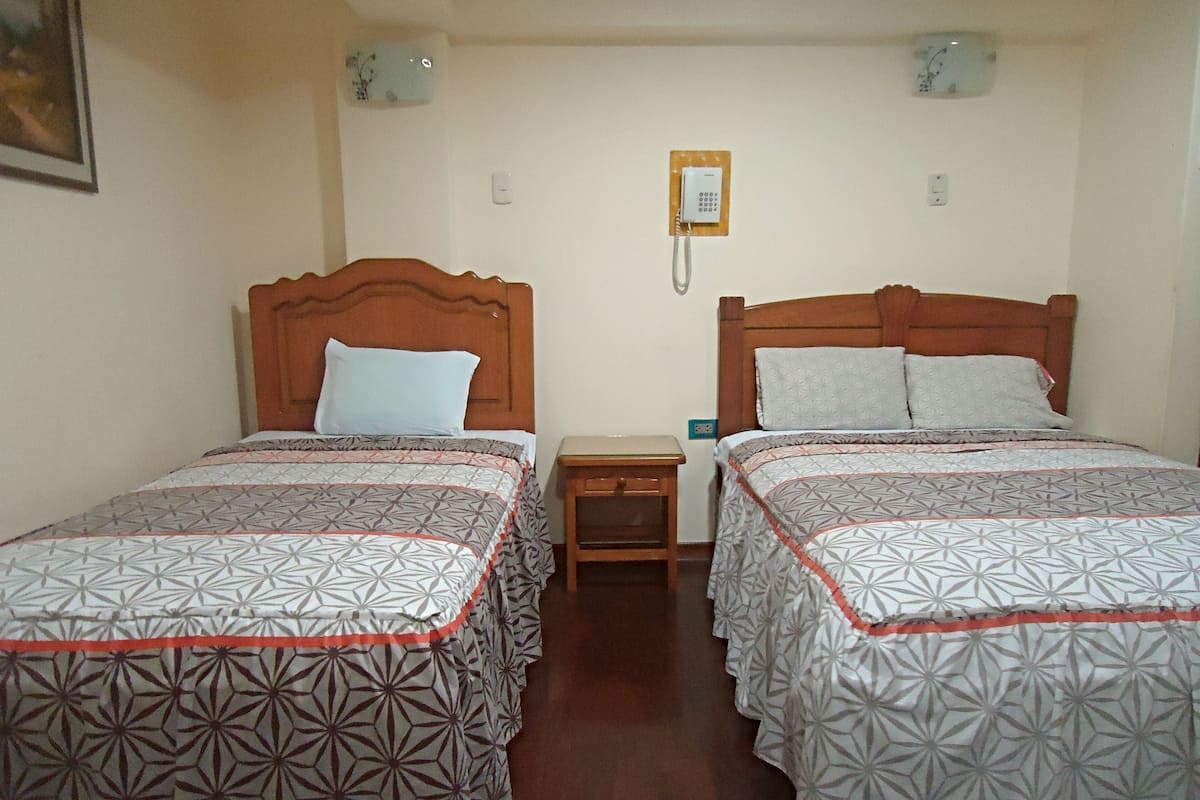 A room with two single beds, each with a wooden headboard and patterned bedding. There is a small wooden nightstand between the beds, and a wall-mounted telephone.