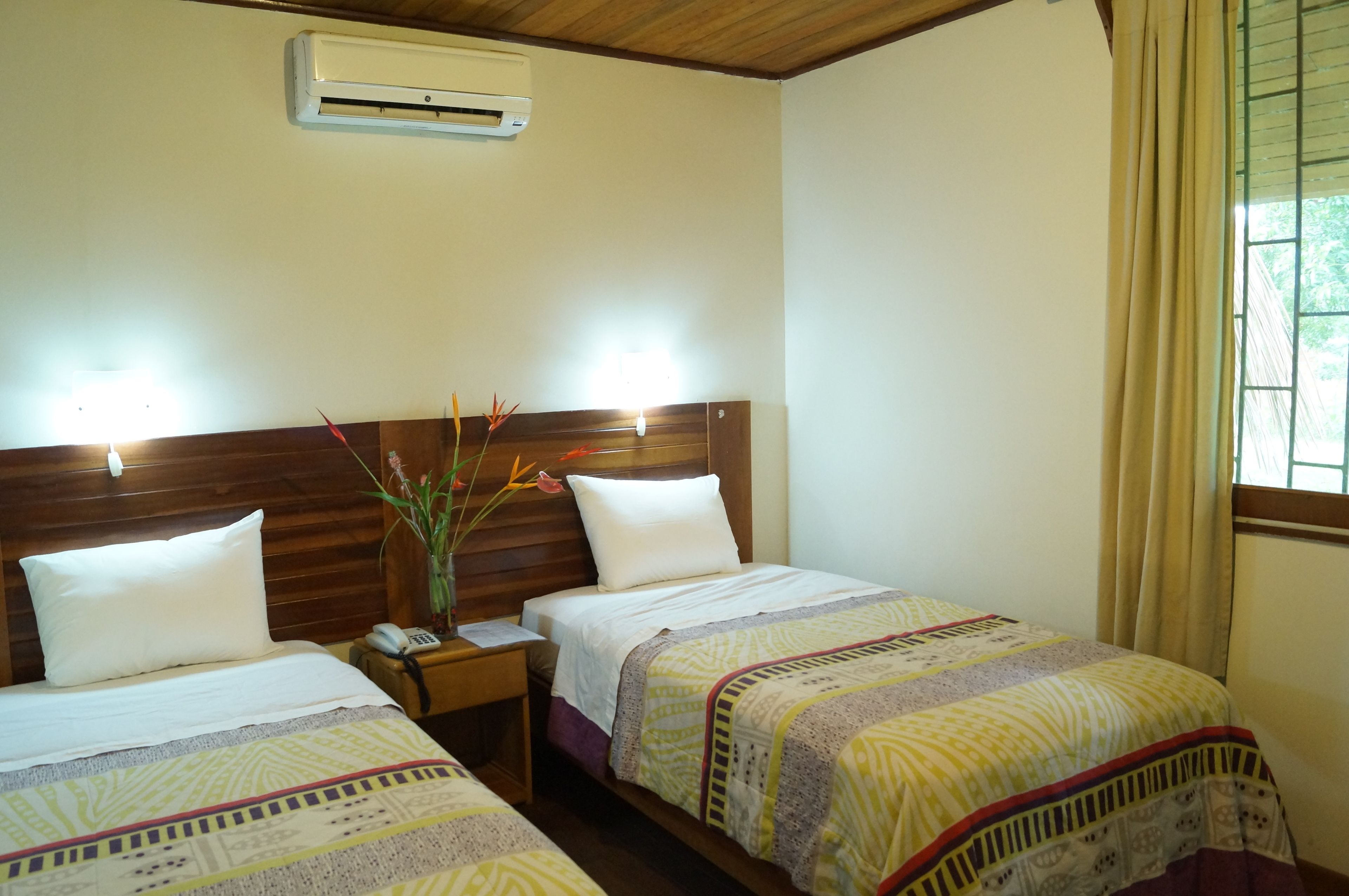 A hotel room with two beds, wooden headboards, a nightstand with a telephone, and an air conditioning unit on the wall.