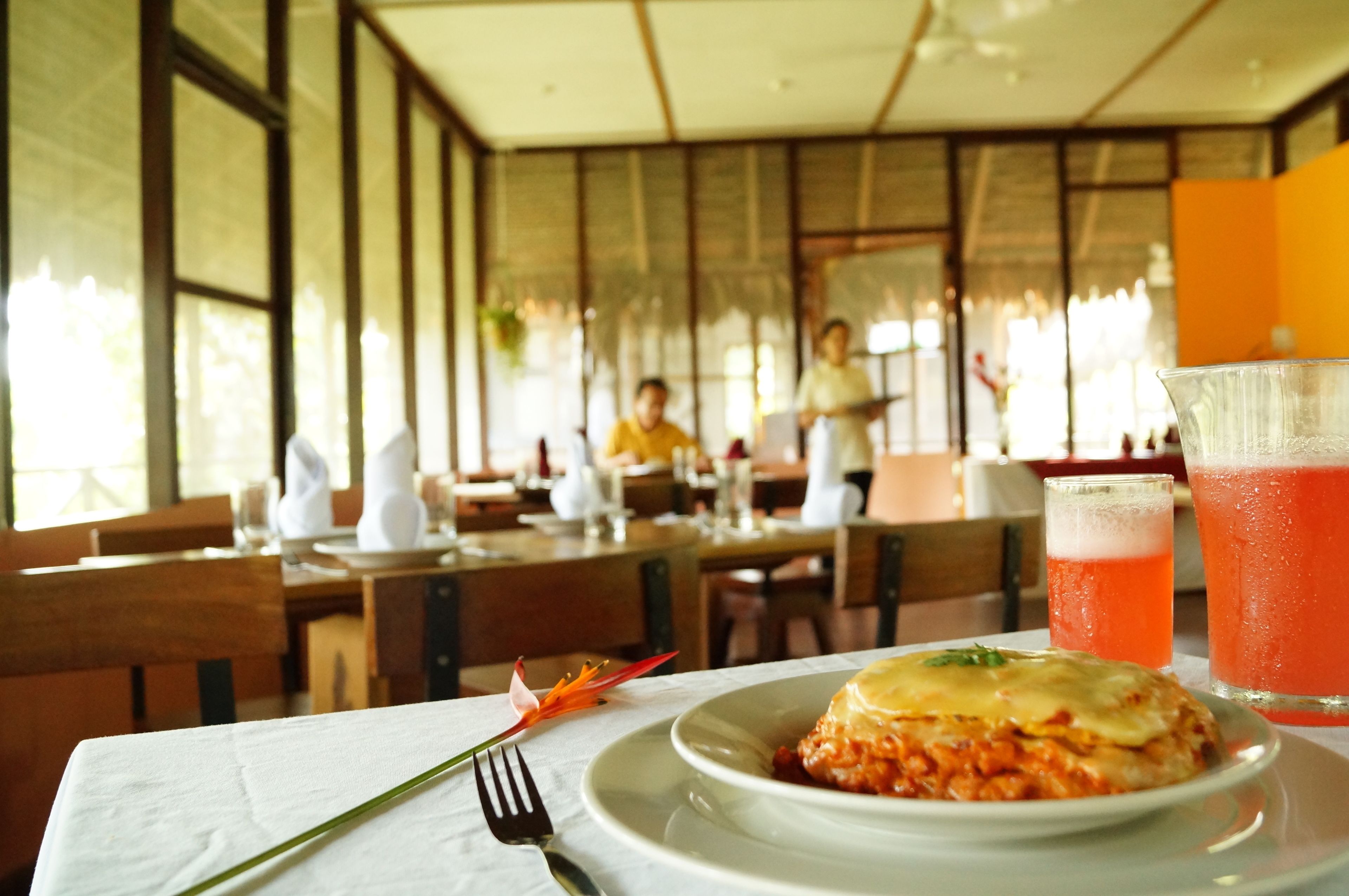 A restaurant interior with wooden tables and chairs, a white tablecloth, and a plate of food with a drink in the foreground.
