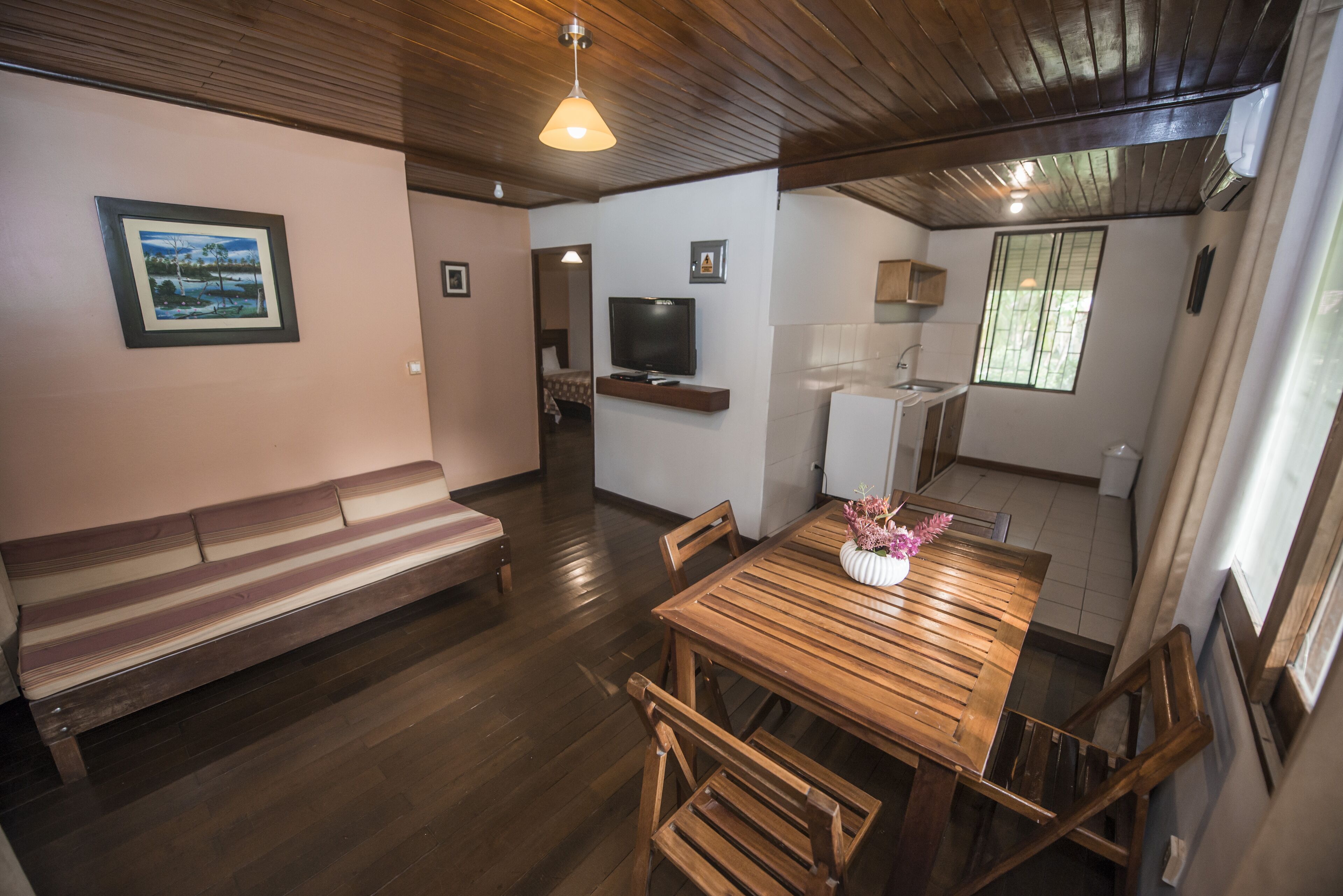 A cozy room with a wooden bench, a dining table with chairs, a TV mounted on the wall, and a window with curtains.