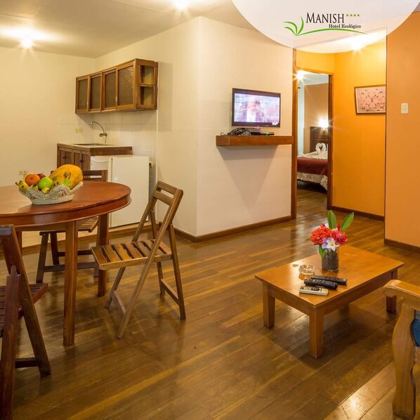A hotel room with a wooden dining table, chairs, a fruit basket, a TV, a bed, and a small table with a vase of flowers. - Manish Hotel Ecológico (Callería)