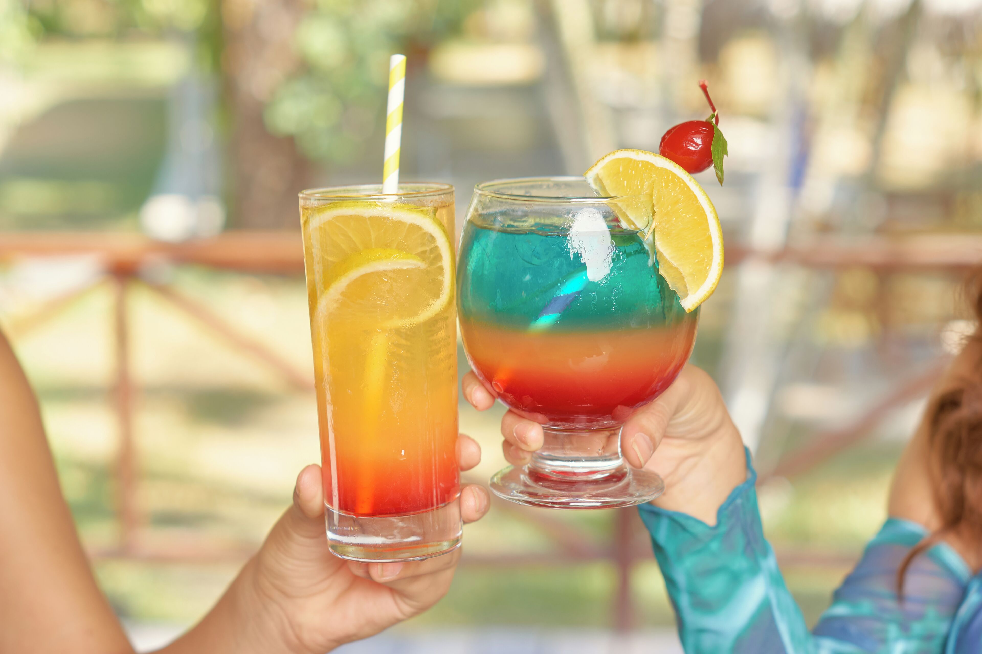 Two people holding glasses of colorful drinks with fruit garnishes.