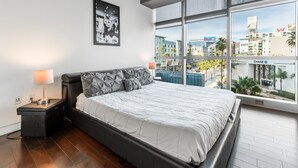 A modern bedroom with a large bed, a nightstand, a lamp, and a city view through large windows.