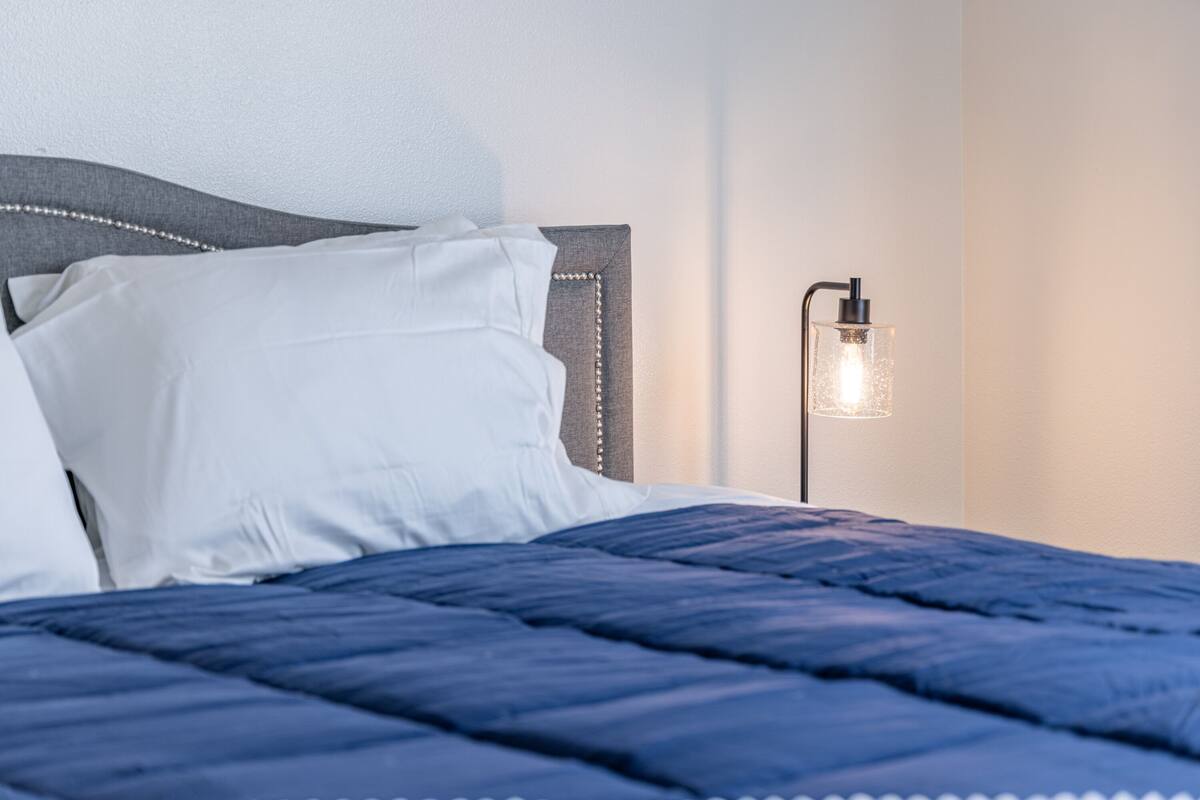 A neatly made bed with a blue quilt, a white pillow, and a headboard with a bead trim. There is a wall-mounted light fixture to the right.
