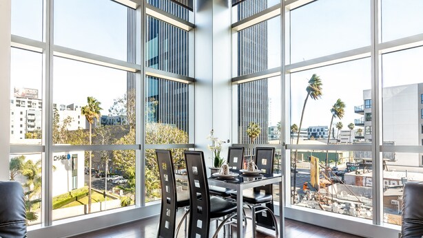 A modern dining area with a large window overlooking a cityscape, featuring a table with chairs and a view of buildings and palm trees.