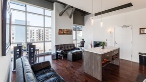 A modern living room with a large window, a black leather sofa, a coffee table, and a small kitchen area with a counter and cabinets.
