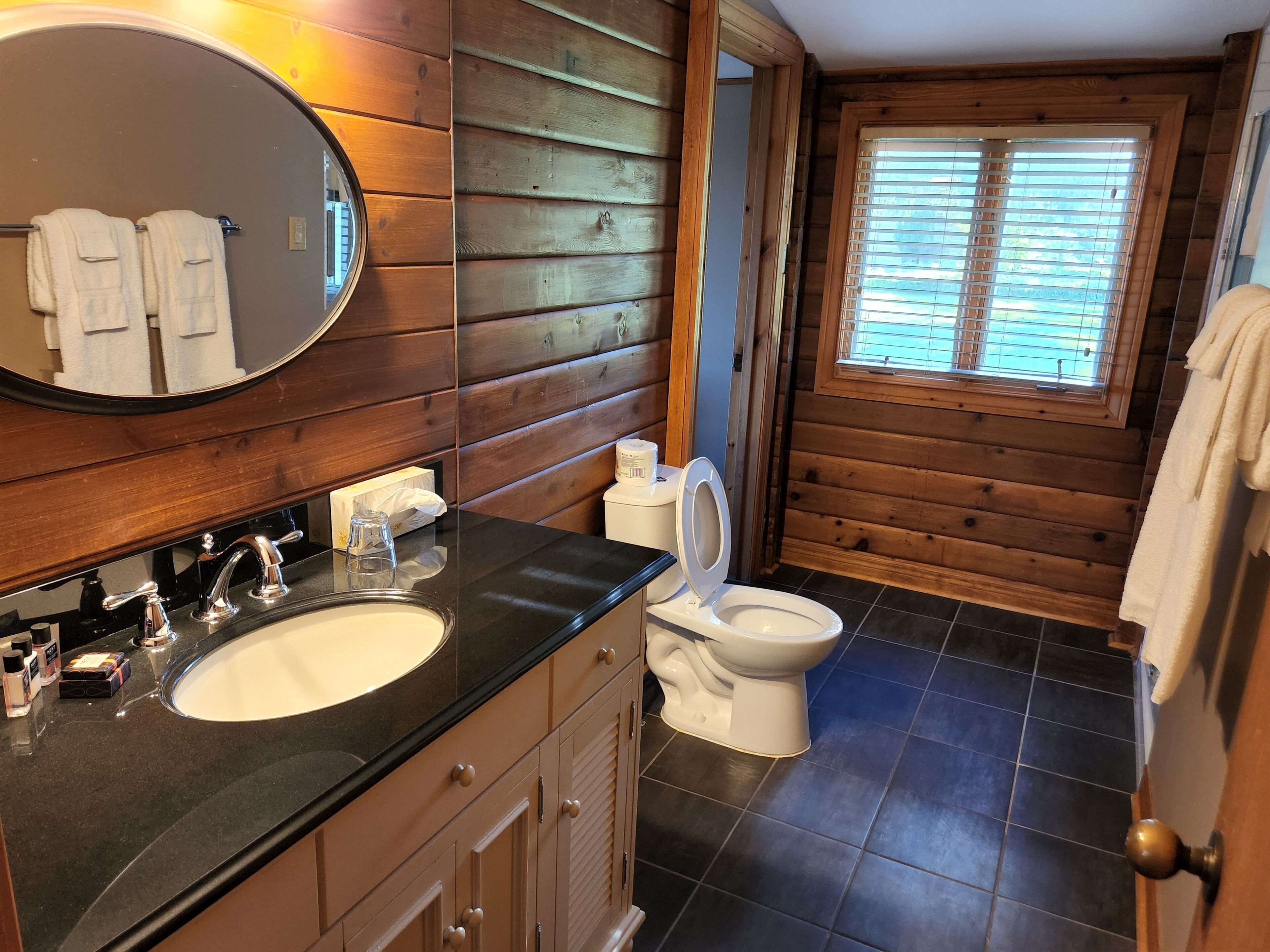 Un baño con paneles de madera, un lavabo con espejo, un inodoro y una ventana con persianas.