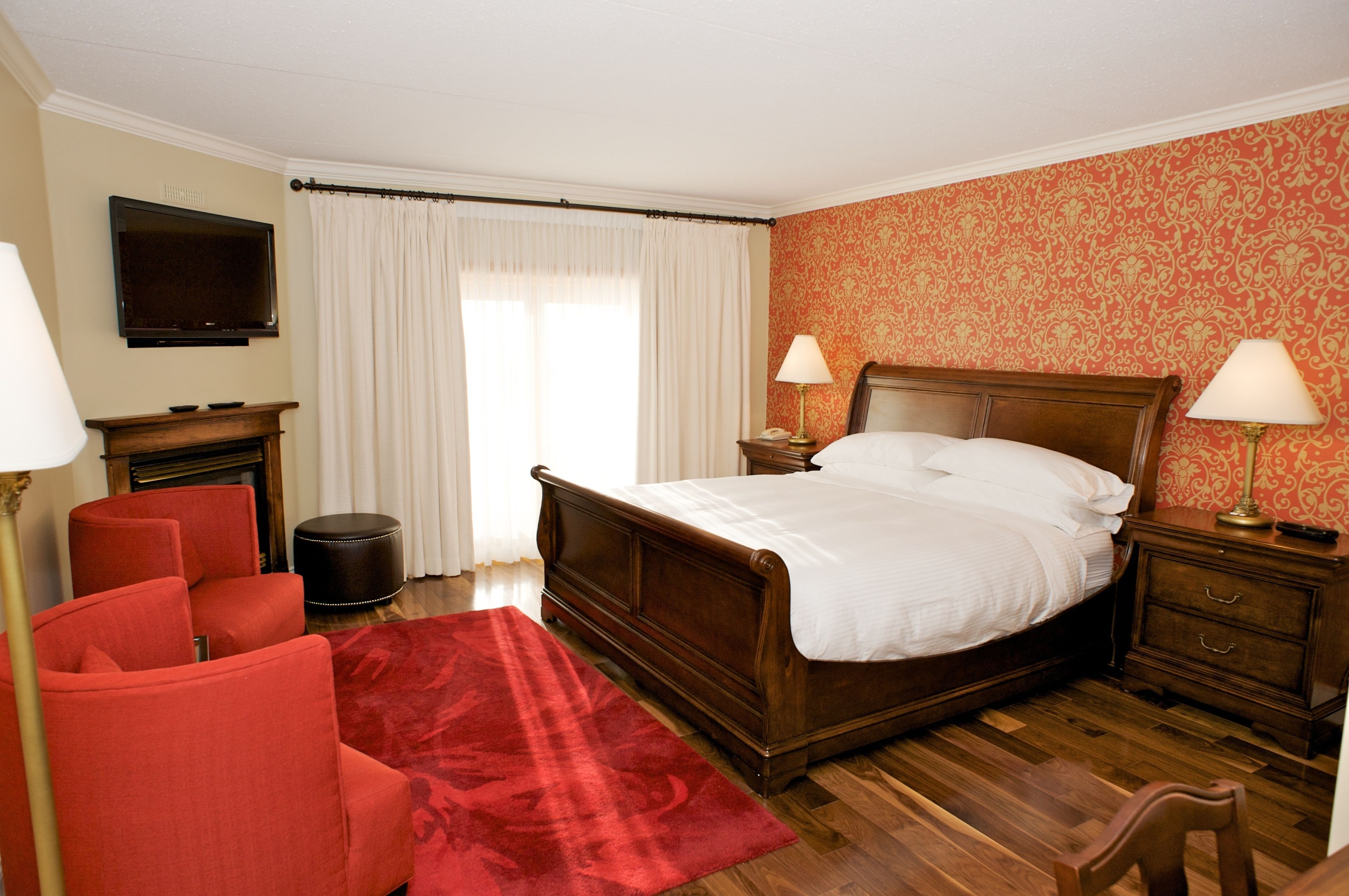 Quarto de hotel com uma cama de madeira, duas luminárias de cabeceira, uma TV fixada na parede, uma poltrona vermelha e um tapete vermelho.
