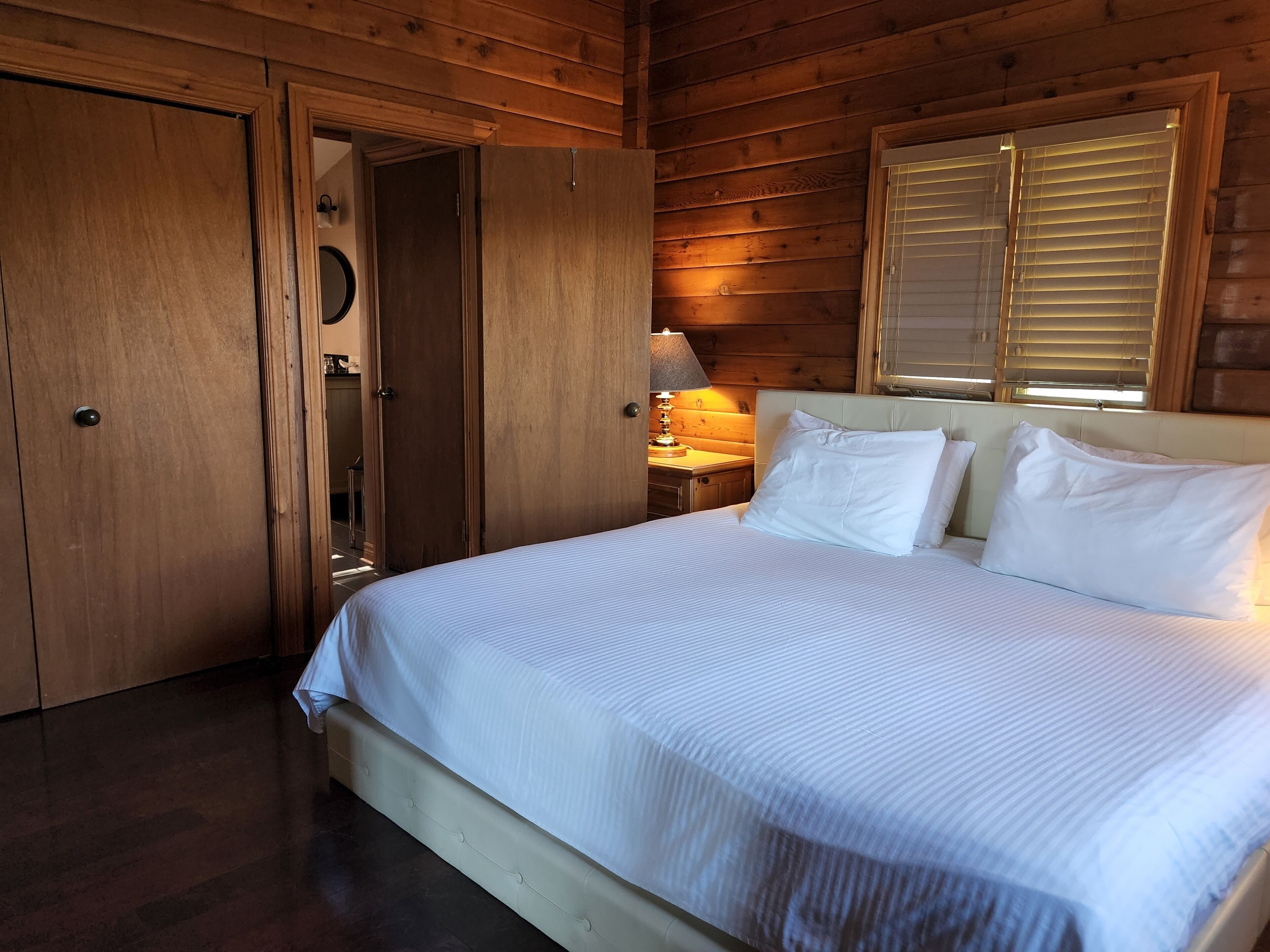 Un dormitorio de cabaña de madera con cama, mesita de noche y ventana con persianas.