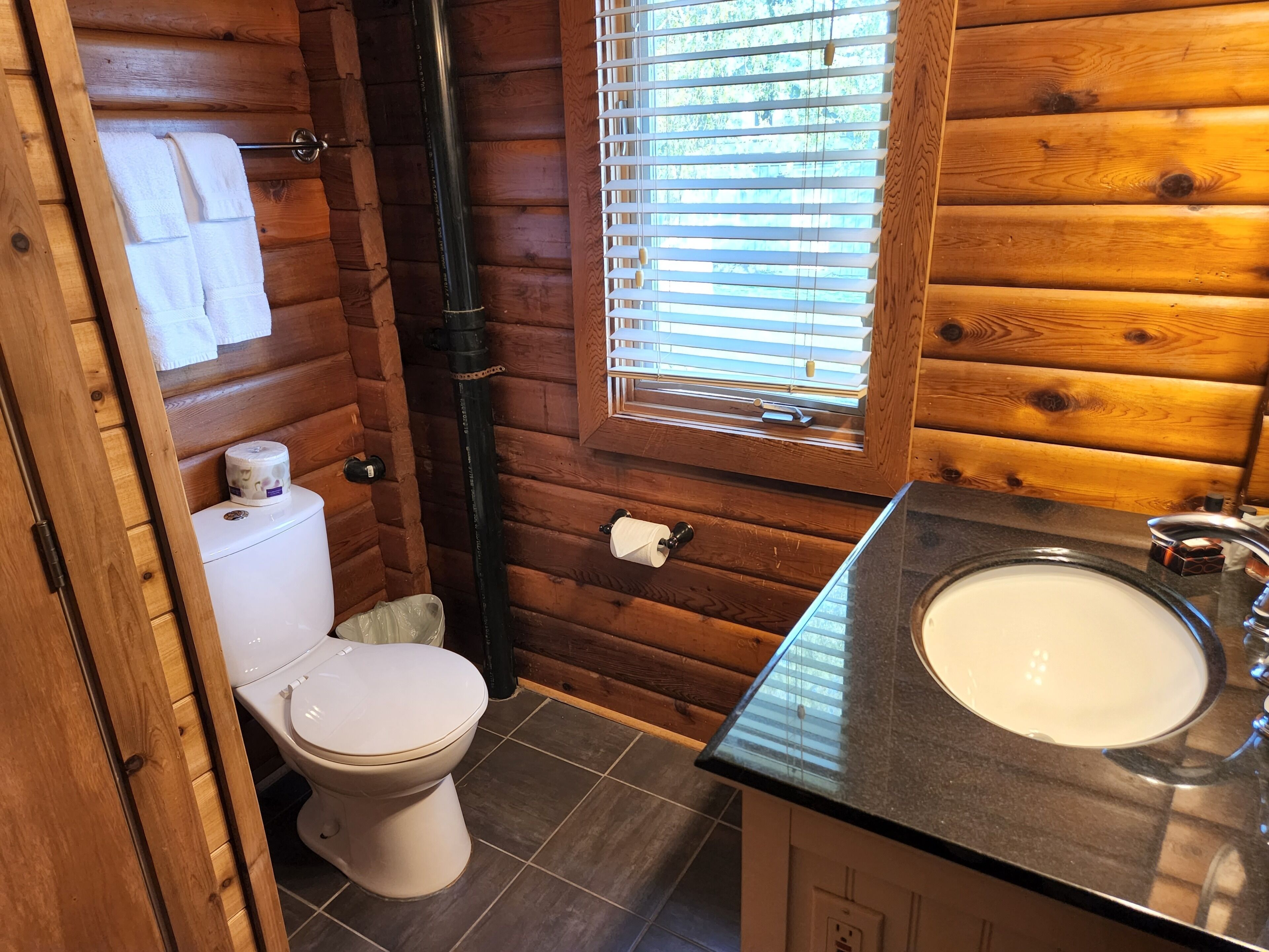 A wooden cabin bathroom with a toilet, a sink, and a window with blinds.