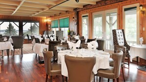 A restaurant with multiple tables set for dining, wooden walls, large windows, and a view of trees.